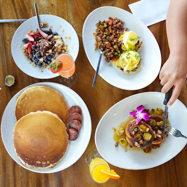 Overhead view of four breakfast plates, including pancakes, eggs Benedict, and fruit, on a wooden table.
